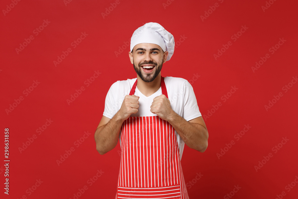 Joyful young bearded male chef cook or baker man in striped apron white t-shirt toque chefs hat posing isolated on red background in studio. Cooking food concept. Mock up copy space. Clenching fists. Joyful young bearded male chef cook or baker man in striped apron white t-shirt toque chefs hat posing isolated on red background in studio. Cooking food concept. Mock up copy space. Clenching fists.