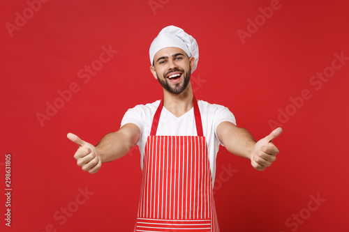 Joyful young bearded male chef cook or baker man in striped apron white t-shirt toque chefs hat posing isolated on red wall background. Cooking food concept. Mock up copy space. Showing thumbs up.