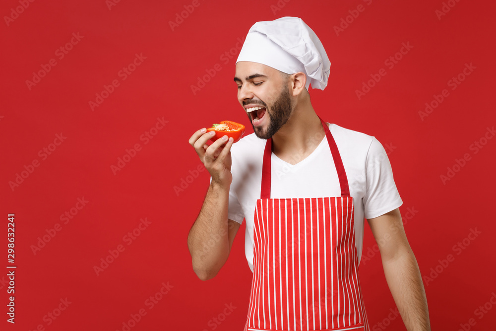 Young bearded male chef cook or baker man in striped apron white t-shirt toque chefs hat isolated red background. Cooking food concept. Mock up copy space. Holding fresh vegetable, biting bell pepper Young bearded male chef cook or baker man in striped apron white t-shirt toque chefs hat isolated red background. Cooking food concept. Mock up copy space. Holding fresh vegetable, biting bell pepper
