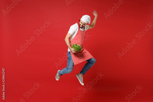 Funny young chef cook or baker man in striped apron toque chefs hat isolated on red background. Cooking food concept. Mock up copy space. Jumping, holding green fresh salad leaves, fooling around.