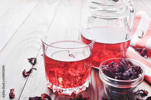 Closeup view at glass of hibiscus ice tea and jug on wooden table background