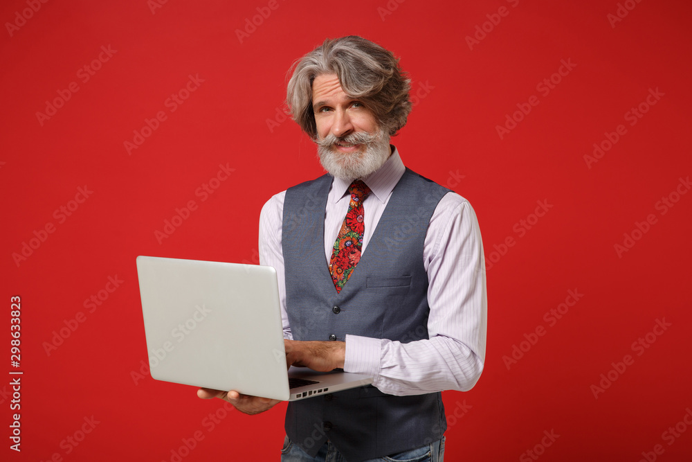 Smiling elderly gray-haired mustache bearded man in classic shirt vest colorful tie posing isolated on red wall background. People lifestyle concept. Mock up copy space. Holding laptop pc computer. Smiling elderly gray-haired mustache bearded man in classic shirt vest colorful tie posing isolated on red wall background. People lifestyle concept. Mock up copy space. Holding laptop pc computer.