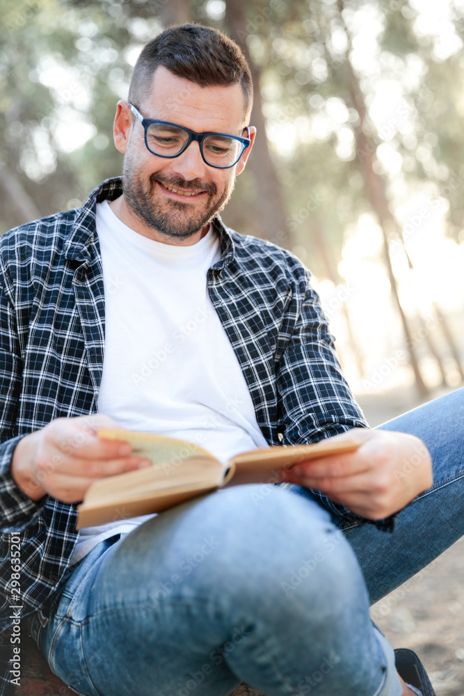 Fototapeta premium Hombre joven con lentes y camisa de cuadros apoyado en un árbol leyendo sonriente un libro al aire libre