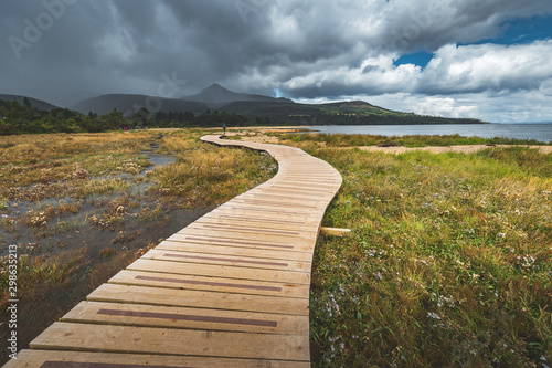 Wooden Walking Way at Coastline Nothern Ireland. Wood Footpath Along Coast  Calm Ocean Bay  Green Forest Climbs and Sky with Stormy Clouds. Wonderful Seaside Place and Tourist Walkway