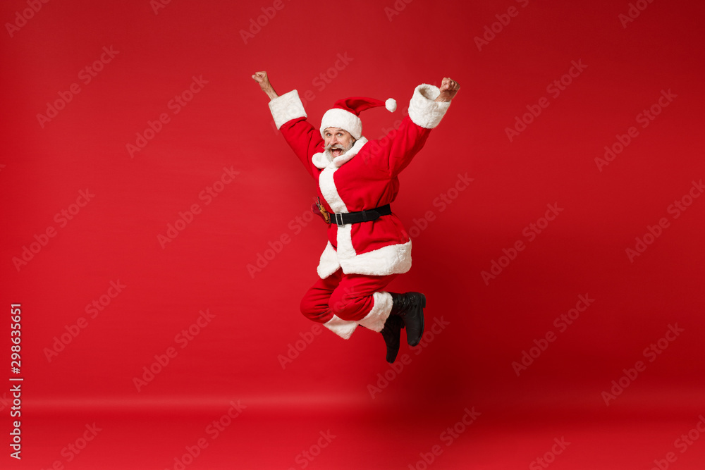 Funny elderly gray-haired mustache bearded Santa man in Christmas hat posing isolated on red wall background. Happy New Year 2020 celebration holiday concept. Mock up copy space. Jumping, having fun. Funny elderly gray-haired mustache bearded Santa man in Christmas hat posing isolated on red wall background. Happy New Year 2020 celebration holiday concept. Mock up copy space. Jumping, having fun.
