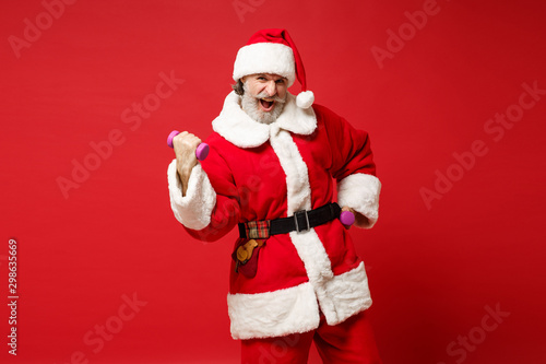 Strong crazy elderly gray-haired mustache bearded Santa man in Christmas hat posing isolated on red background. Happy New Year 2020 celebration holiday concept. Mock up copy space. Holding dumbbells.