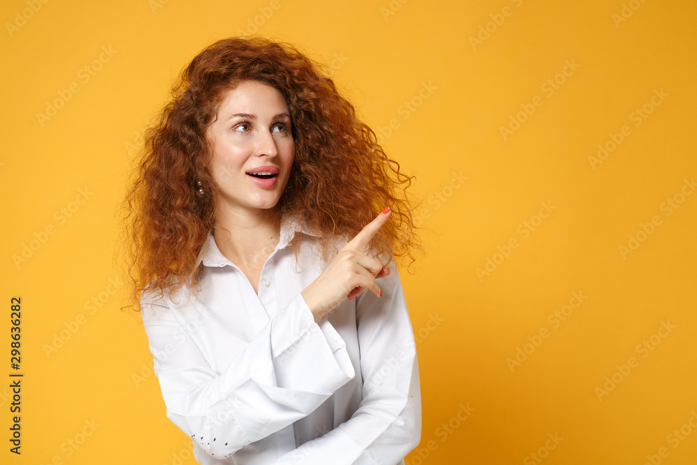 Amazed young redhead woman girl in white shirt posing isolated on yellow orange background studio portrait. People sincere emotions lifestyle concept. Mock up copy space. Pointing index finger aside. Amazed young redhead woman girl in white shirt posing isolated on yellow orange background studio portrait. People sincere emotions lifestyle concept. Mock up copy space. Pointing index finger aside.