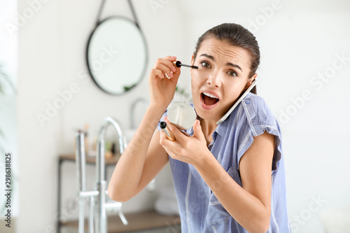 Fototapeta Naklejka Na Ścianę i Meble -  Hurrying woman applying makeup while talking by phone in morning