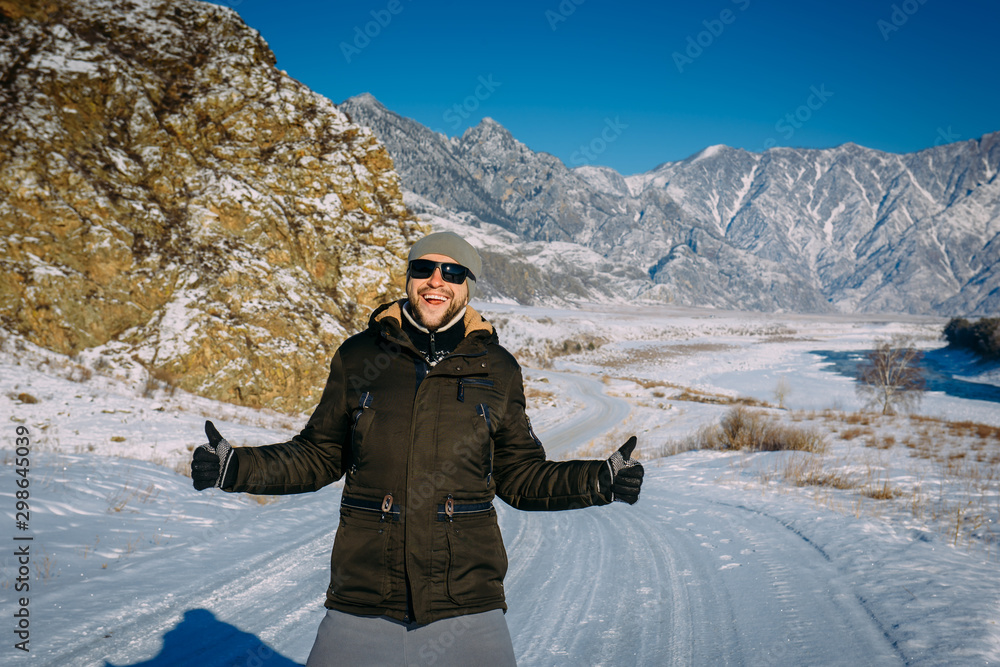Young cheerful guy enjoying Christmas vacation in mountains. Attractive man  laughs and gives thumbs up against snow-capped mountains and blue sky. New year's journey, active winter tourism concept.