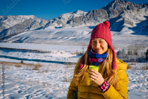 Beautiful girl in red hat holds glass on the background of snow-capped mountains on frosty sunny day. Cheerful young woman enjoying Christmas vacation in the mountains, looking at camera and smiling.