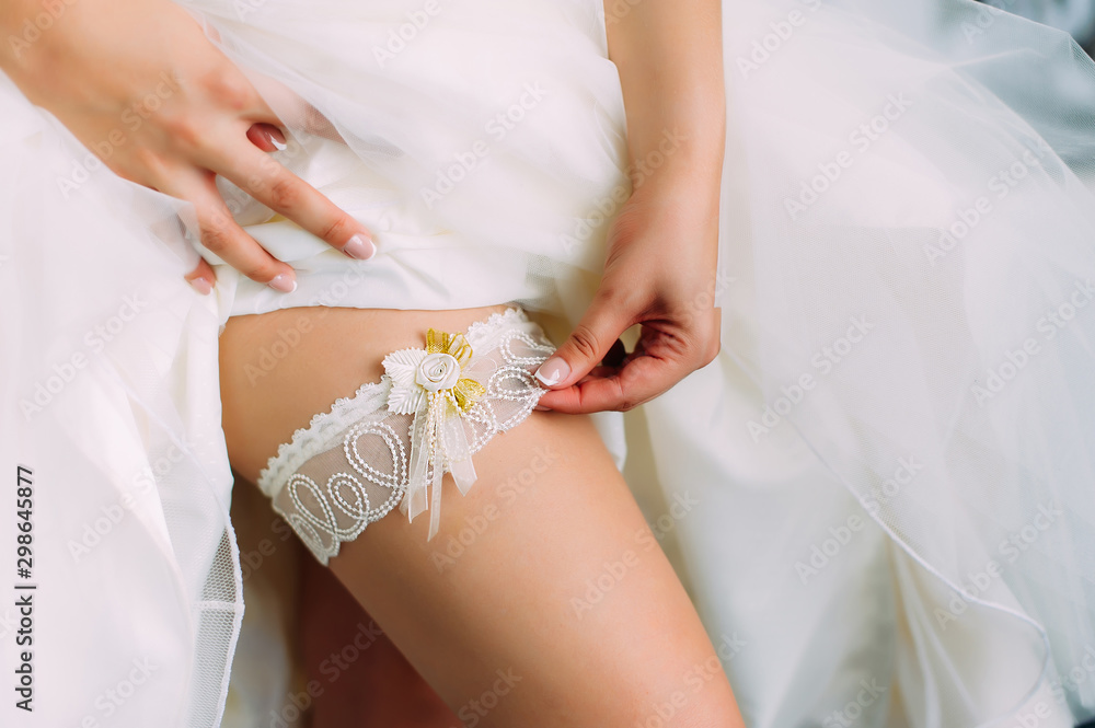 Young bride in a white dress with a wedding garter on her leg, close-up. Preparation for the wedding celebration. Accessories for the newlyweds, lace garter. Young bride in a white dress with a wedding garter on her leg, close-up. Preparation for the wedding celebration. Accessories for the newlyweds, lace garter.