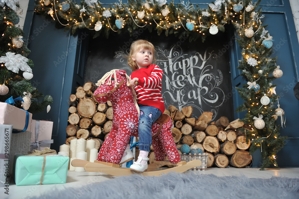 Little girl child sitting on a red rocking horse and smiling on the background of the Christmas interior with a fireplace, firewood and gifts