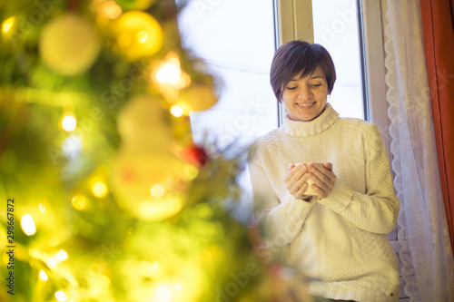 Young beautiful woman sitting home by the window with cup of hot coffee wearing knitted warm sweater.