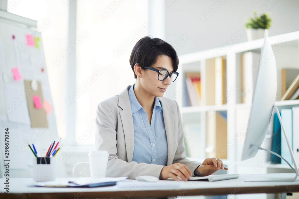 Side view portrait of young businesswoman using computer while sitting at desk in office, copy space Side view portrait of young businesswoman using computer while sitting at desk in office, copy space