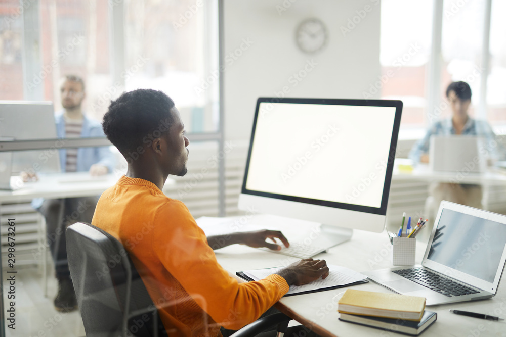 © Seventyfour - Side view portrait of young African-American man using computer in office, focus on blank white screen, copy space