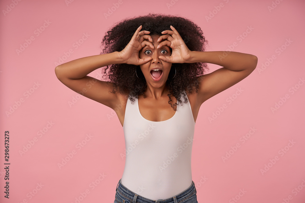 Cheerful young curly female with casual hairstyle wearing white shirt and jeans, making fun over pink background, looking at camera happily and folding eyeglasses with her hands