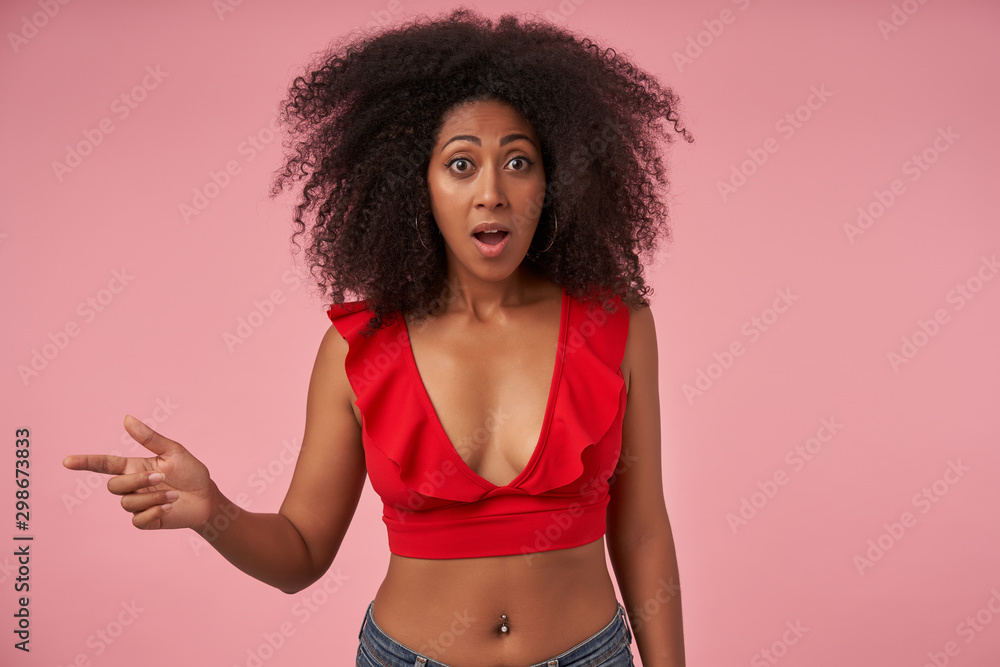 Young bemused curly dark skinned woman with belly button piercing pointing  aside with forefinger while posing over pink background, looking to camera  with wde eyes and mouth opened Stock Photo | Adobe