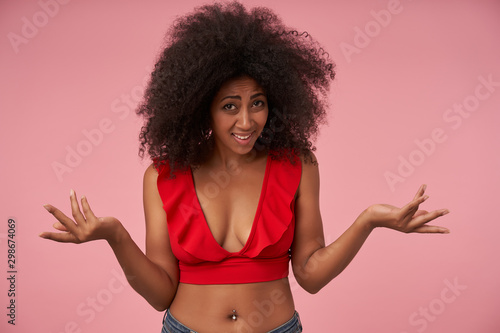 Indoor photo of dissatisfied young dark skinned lady with casual hairstyle wearing red top and jeans, raising palms perplexedly and contracting forehead, isolated over pink background photo