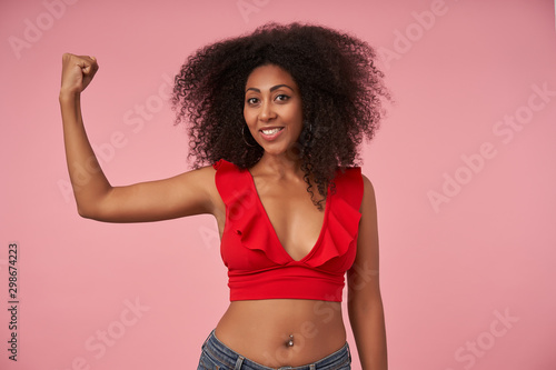 Positive pretty curly dark skinned female with belly button piercing looking at camera with wide cheerful smile, demonstrating her power in raised hand, posing over pink background