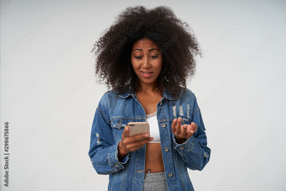 Young curly woman with dark skin holding mobile phone in hand and looking at screen wonderingly, wearing white top and jeans coat, posing over white background Young curly woman with dark skin holding mobile phone in hand and looking at screen wonderingly, wearing white top and jeans coat, posing over white background