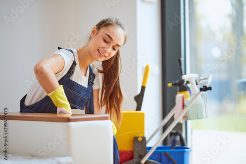 Positive caucasian girl in special equipment for cleaning carefully wiping dust off from white sofa in new house. Detergents background photo