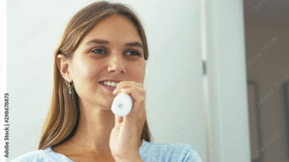 A happy woman in a bright bathroom, washing her teeth with an electric toothbrush. Interior handheld shot, morning, in front of the mirror. A happy woman in a bright bathroom, washing her teeth with an electric toothbrush. Interior handheld shot, morning, in front of the mirror.
