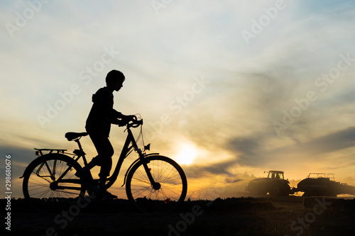 Boy , kid 10 years old riding bike in countryside, tractor working in background, silhouette of riding person and machine at sunset in nature