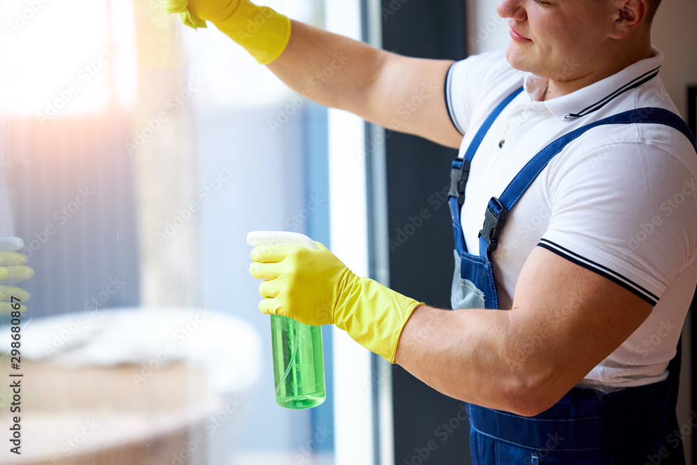 Young muscled guy in uniform from cleaning service wearing yellow protective gloves, holding spray wash the window Young muscled guy in uniform from cleaning service wearing yellow protective gloves, holding spray wash the window