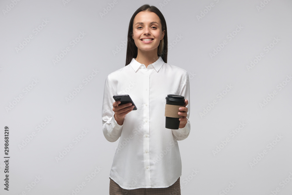 Young European woman in formal clothes, holding phone in one hand and coffee cup in another, isolated on grey background Young European woman in formal clothes, holding phone in one hand and coffee cup in another, isolated on grey background