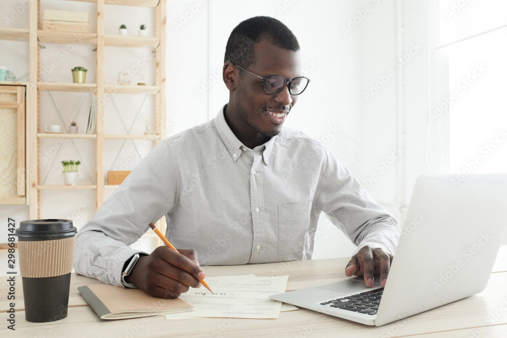 Closeup of African American guy working in office, writing in paper documents and typing data on laptop Closeup of African American guy working in office, writing in paper documents and typing data on laptop