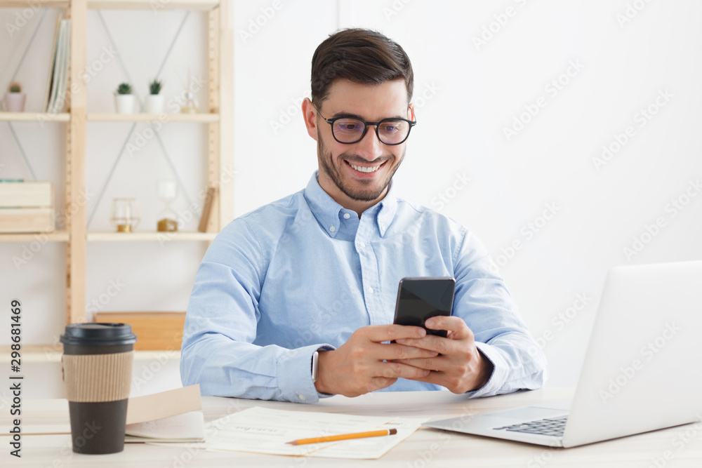 Young male in trendy glasses exchanging messages on smartphone while working in office Young male in trendy glasses exchanging messages on smartphone while working in office