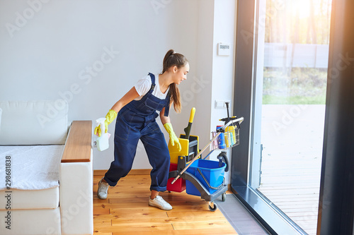 Young caucasian girl with cleaning trolley full of detergents, cleaning room in house with panoramic window. Woman dressed in blue uniform, in process of working photo