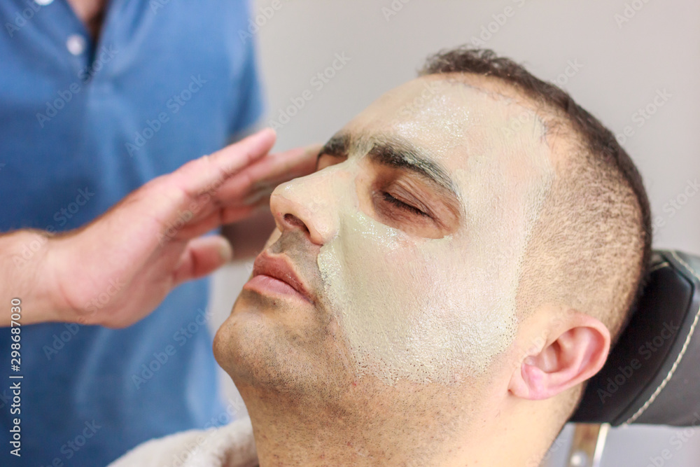 Obraz premium Male skin care in a beauty salon. Applying clay cleansing mask on a man's face
