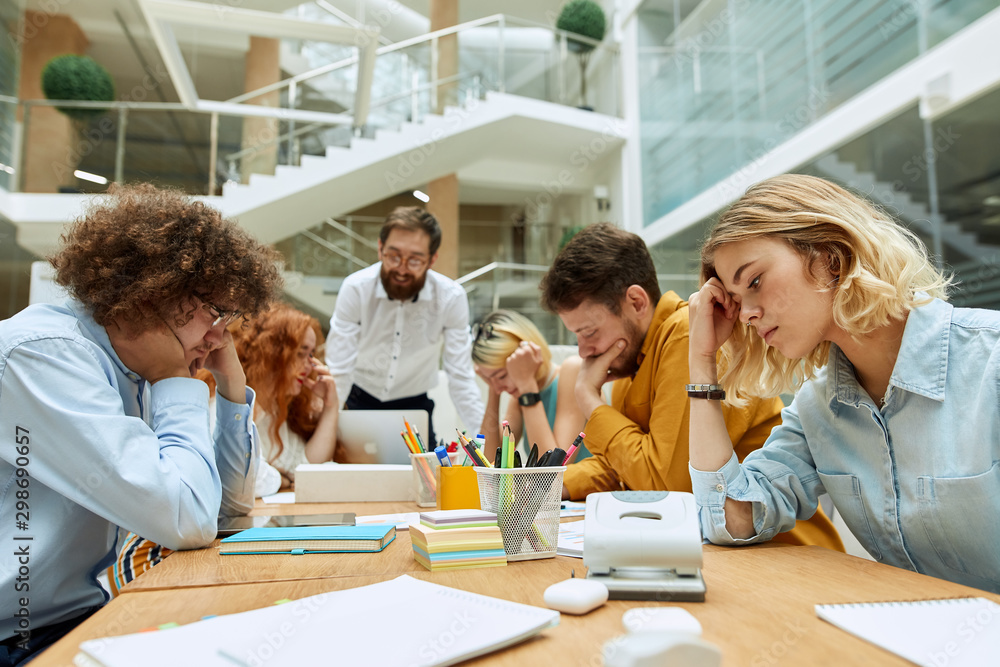 Obraz premium Middle aged creative guy in spectacles with Afro hairstyle, sits opposite young blonde colleague, closes eyes, keeps neck with hands with thoughtful expression in comfortable place