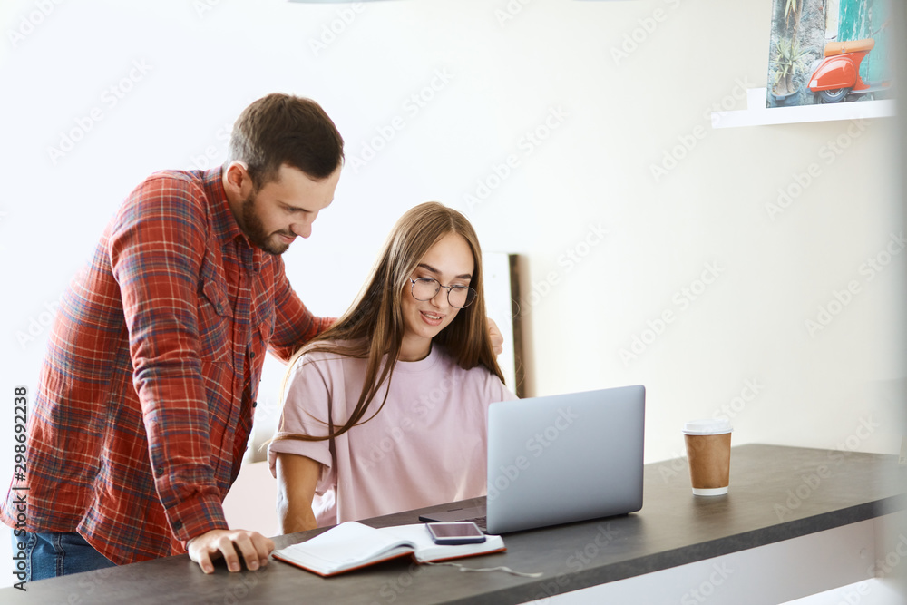 Handsome young man with thick beard in red shirt, stands behind wife, focuses on screen, smiling positively, side shot, family concept Handsome young man with thick beard in red shirt, stands behind wife, focuses on screen, smiling positively, side shot, family concept