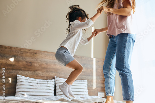 Portrait of little joyful six years old girl happily jumping on bed together with sister in brightly lighted bedroom, shot from below, family joy concept