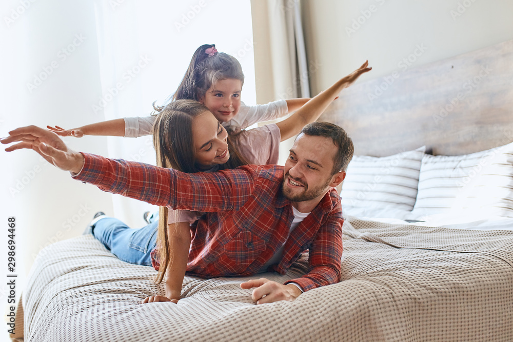 Happy loving family spending time together, father, mother and daughter playing cheerfully on big bed in hotel room, stretching hands, having fun, smiling emotively, expressing positive mood, - obrazy, fototapety, plakaty 