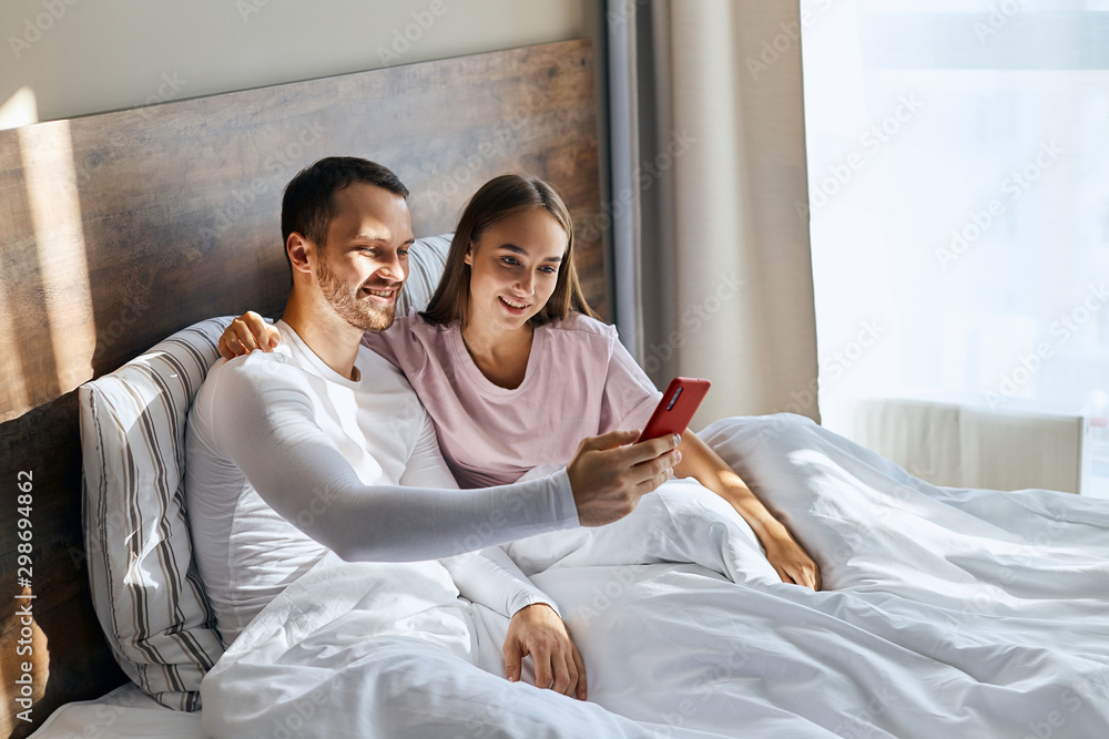 Top view of impressive handsome husband with unshaven face, stretching hand with cell phone ahead, taking selfie together with young wife sitting in bed covered with blanket, love concept Top view of impressive handsome husband with unshaven face, stretching hand with cell phone ahead, taking selfie together with young wife sitting in bed covered with blanket, love concept