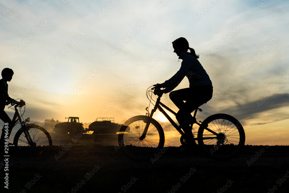 Obraz premium Boy , kid 10 years old, and girl riding bikes in countryside, tractor working in background, silhouette of riding persons and machine at sunset in nature