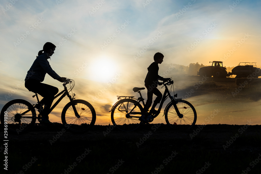 Obraz premium Boy , kid 10 years old, and girl riding bikes in countryside, tractor working in background, silhouette of riding persons and machine at sunset in nature