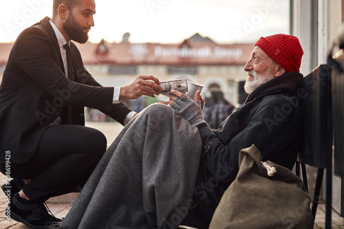 Decent rich man in tuxedo hunkered down to homeless and give money donation, one dollar bill to beggar male photo