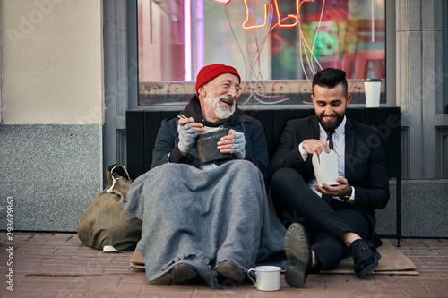 Stylish office worker and beggar male sit eating on street and speaking. Man un tuxedo, poor male in old clothes photo