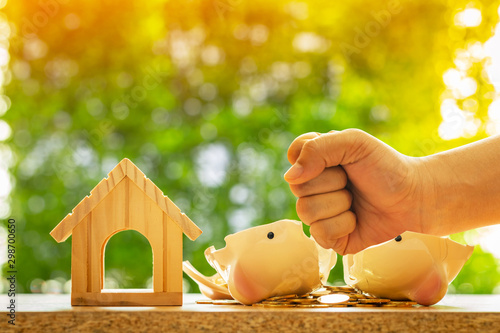 Man hand over which is raised above broken piggy bank with money and wooden home model on bokeh background in the public park, Loan and debt of close account of business real estate concept