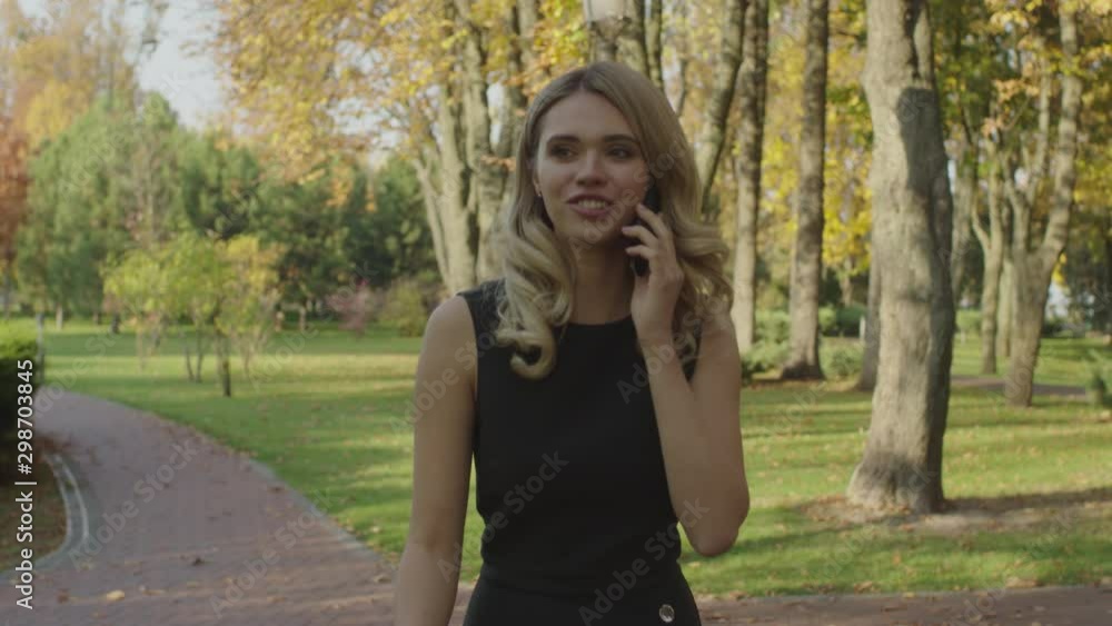 A blonde smiling girl dynamically walks along a park alley and speaks on the phone A blonde smiling girl dynamically walks along a park alley and speaks on the phone