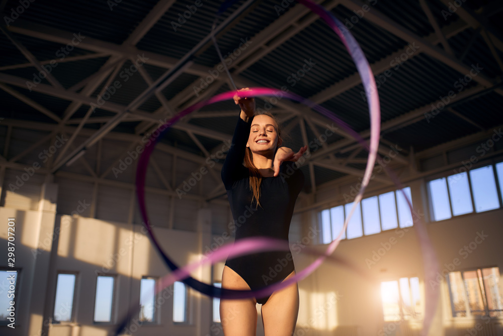 Charming young girl with closed eyes and bright smile dancing with violet ribbon in sports hall, portrait, close up, rhythmic gymnastics concept