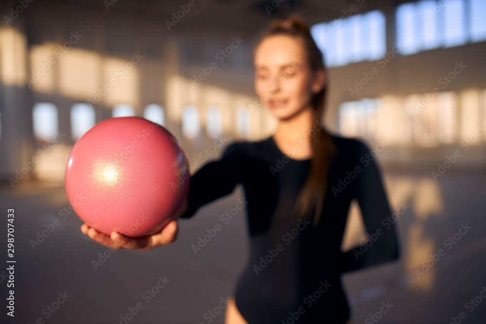 Young female gymnast dressed in black leotard stretching hand with pink ball towards camera, blurred background, indoor shot