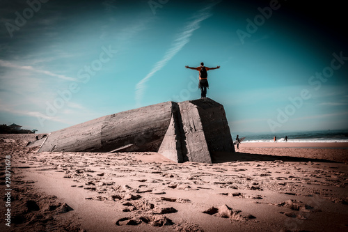 Labenne Océan, les Landes / France »; October 26, 2019: A young man with open arms climbed a Bunkers on the beach of Labenne Océan, with ocher retouching photo