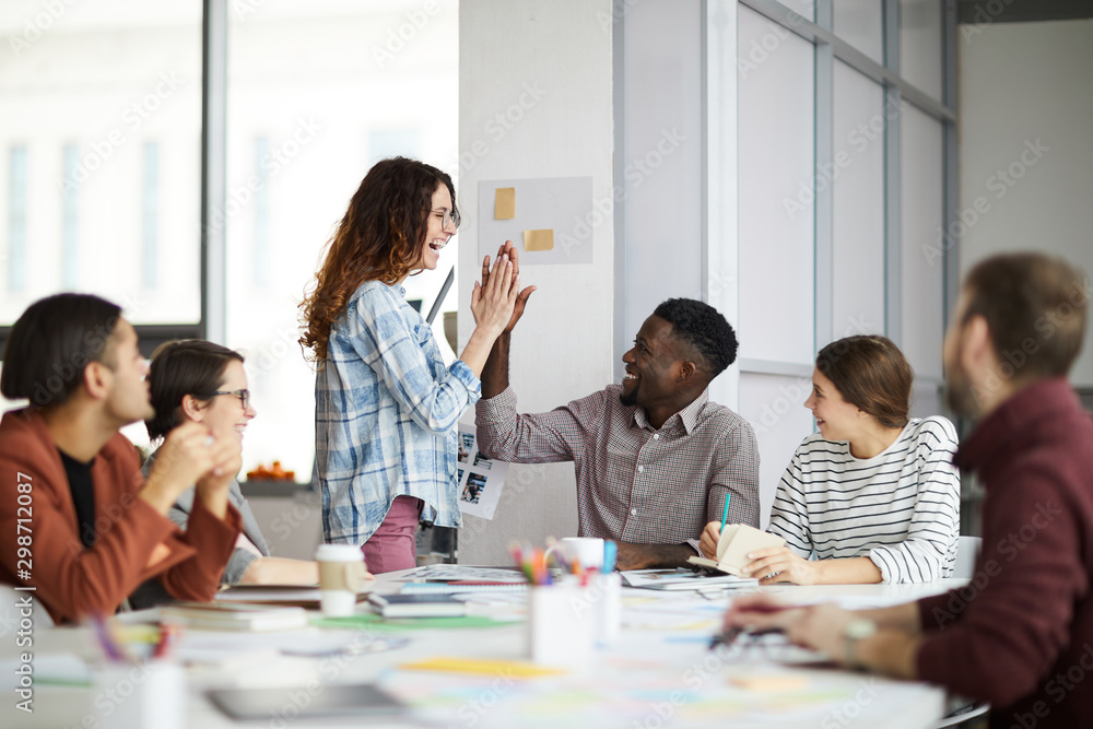 Fototapeta premium Portrait of creative business team giving high five while planning project during meeting in office, focus on smiling young woman standing by table