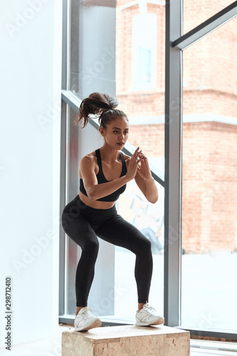 Jumping on fit box in process. Young athletic woman do squats on wooden box in gym. Window background photo