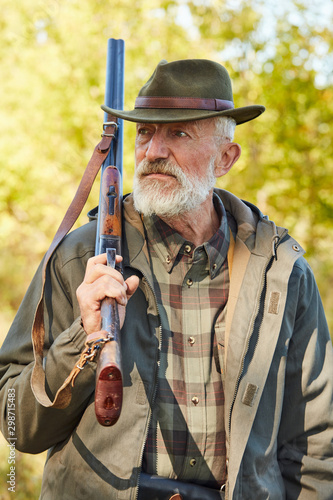 Senior caucasian man with hunting gun looking away, man with grey beard wearing hunting clothes outdoors. Forest background